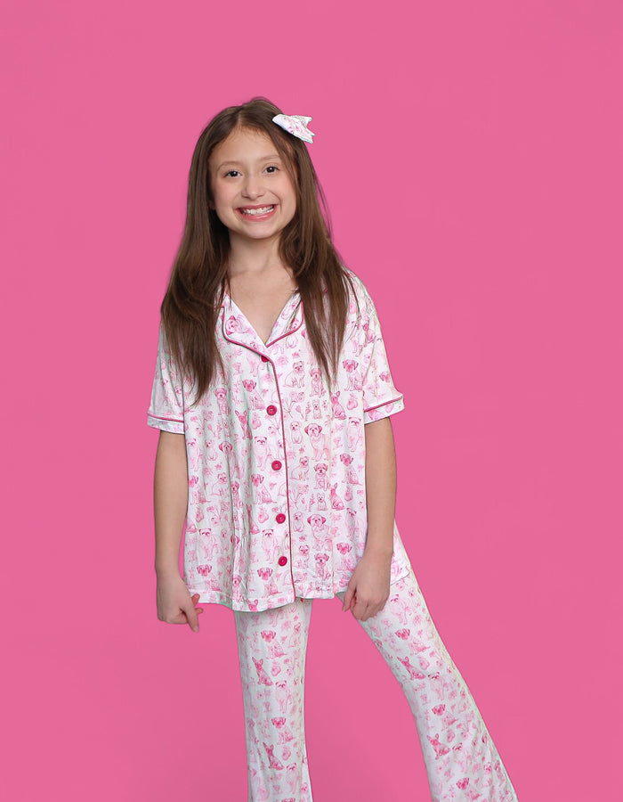 Girl wearing white short-sleeve pajama top and flare pants with pink puppy toile print and button front.
