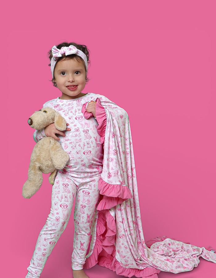 Child wearing pink and white puppy print pajama set with matching headband and ruffled cape, holding stuffed dog toy.