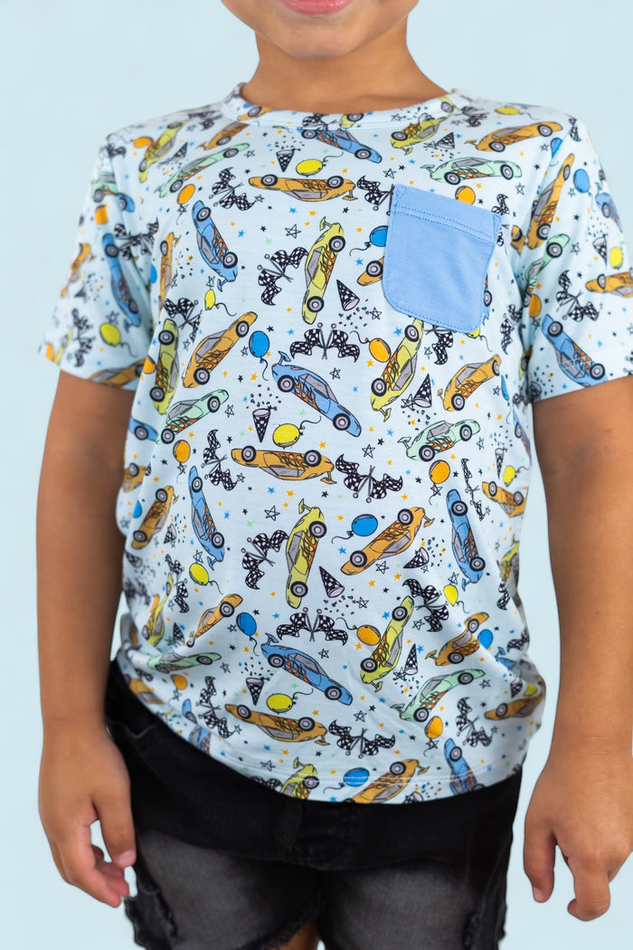 Light blue short-sleeve tee with racing cars, balloons, and stars print, featuring a blue chest pocket.
