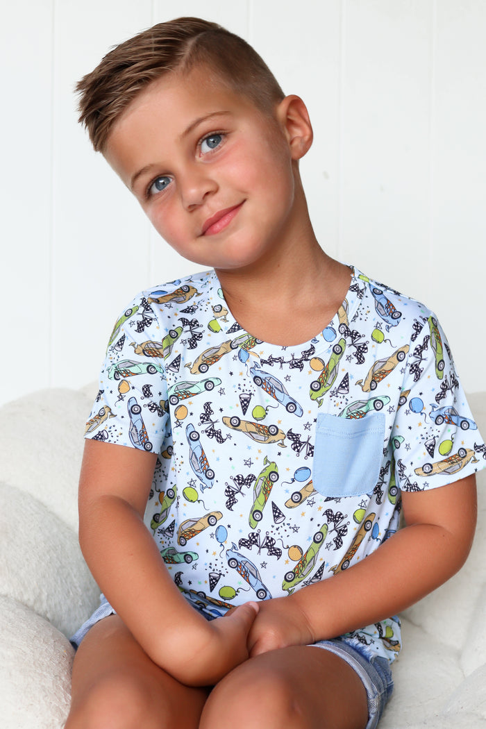 Child wearing light blue pocket tee with racing cars, rockets, and stars printed throughout.