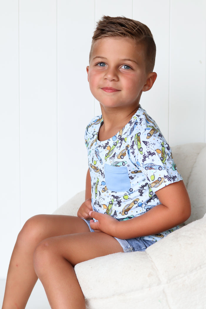 Boy wearing white short-sleeve pocket tee with racing car print in blue, yellow, and purple tones.