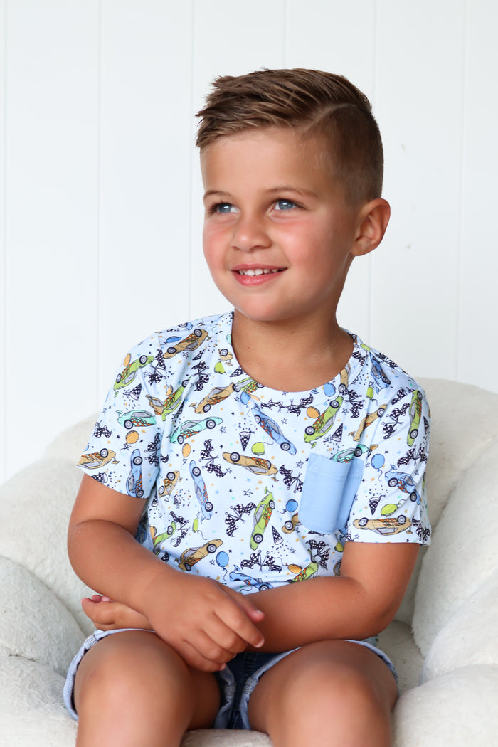 Light blue short-sleeve pocket tee with racing cars, planes, and balloons print worn by young boy.