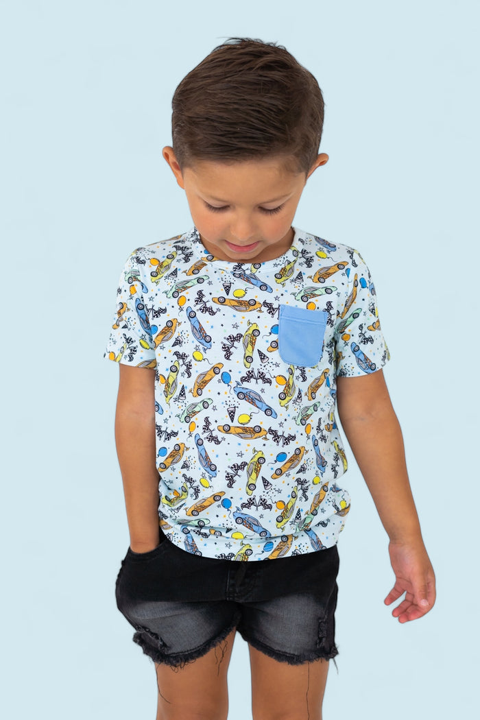 White short-sleeve tee with racing cars, planets, and stars print; blue chest pocket; worn with black distressed denim shorts