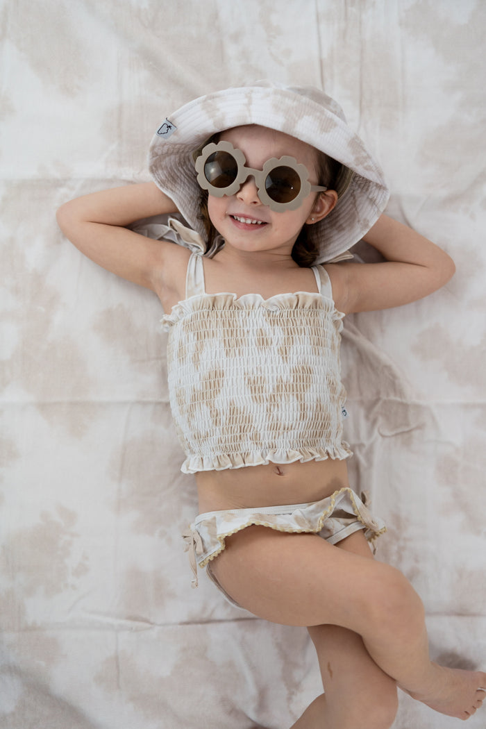 Child lying on beige and white striped towel wearing matching two-piece swimsuit and wide-brimmed hat.