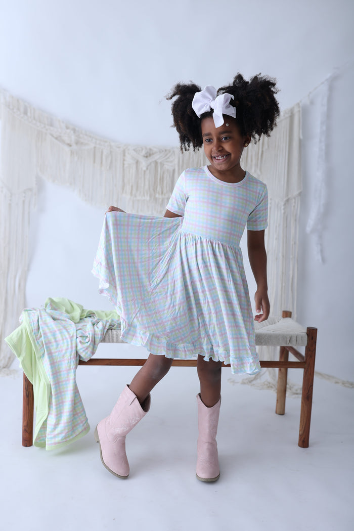 Girl wearing short-sleeve gingham dress in pastel pink and blue with ruffled hem, paired with pink boots.