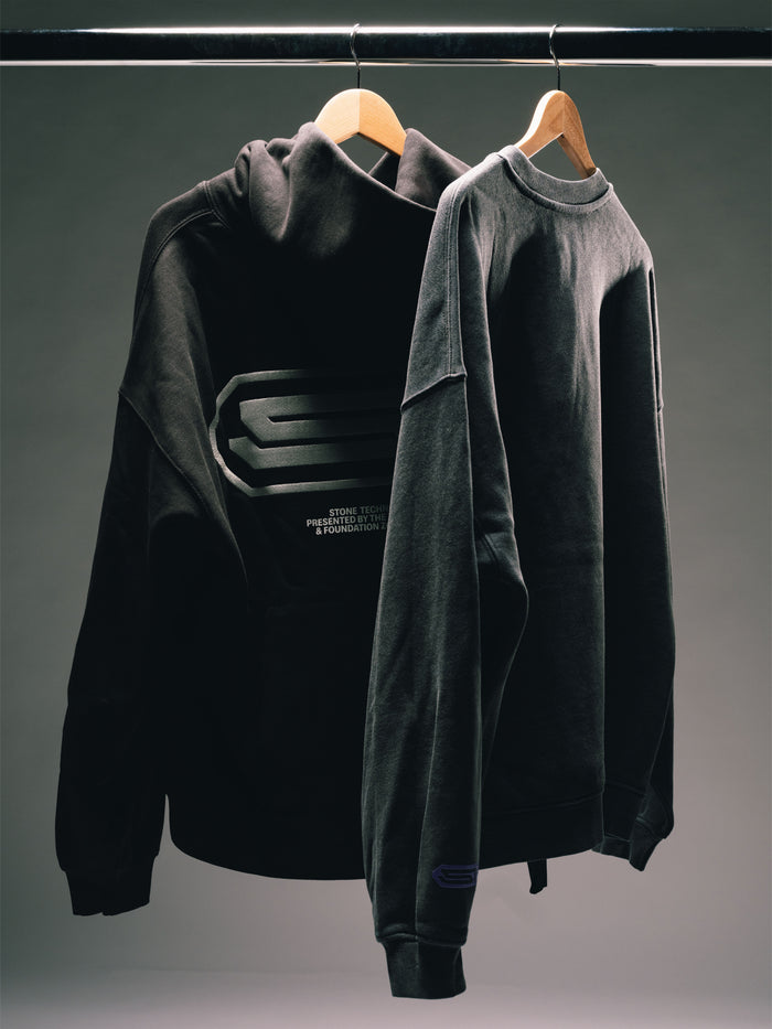 Schwarzer Hoodie und dunkelgraues Sweatshirt auf Holzbügeln vor grauem Hintergrund, mit dezenten Logo- und Printdetails.