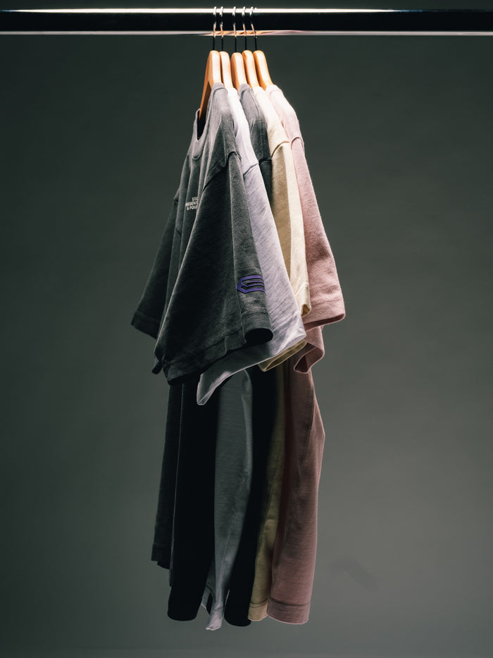Mehrere T-Shirts in Schwarz, Grau, Beige und Rosa dicht nebeneinander auf Holzbügeln an einer schwarzen Kleiderstange.
