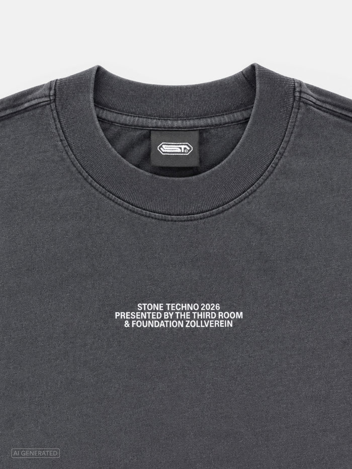 Nahaufnahme des dunkelgrauen T-Shirts mit weißem Brustprint „STONE TECHNO 2026 PRESENTED BY THE THIRD ROOM & FOUNDATION ZOLLVEREIN“. Sichtbar sind außerdem der gerippte Rundhalskragen und das schwarze Nackenlabel mit Stone-Techno-Logo.