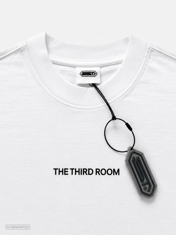 Nahaufnahme des weißen T-Shirts mit schwarzem Schriftzug „THE THIRD ROOM“ auf der Brust. Sichtbar sind außerdem der gerippte Rundhalskragen, das weiße Nackenlabel mit Logo sowie ein grauer Logo-Anhänger am Hangtag.